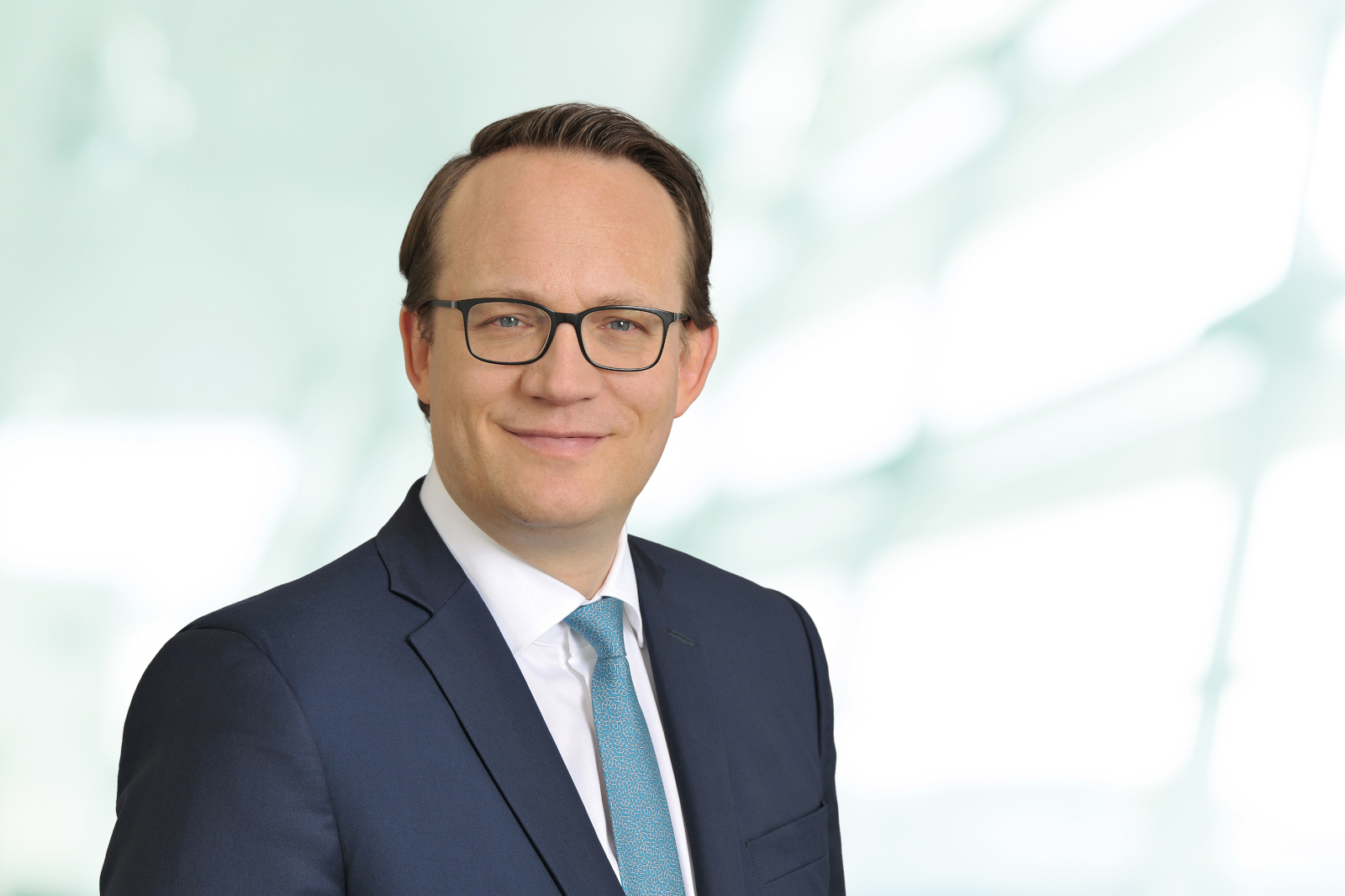 Ein Mann in einem dunklen Anzug mit einer blauen Krawatte steht vor einem unscharfen, hellen Hintergrund.